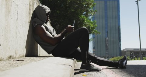 Side view of an African American man wearing a sleeveless hoodie and sportswear sitting on a step using a smartphone before training on a sunny day in the city, slow motion