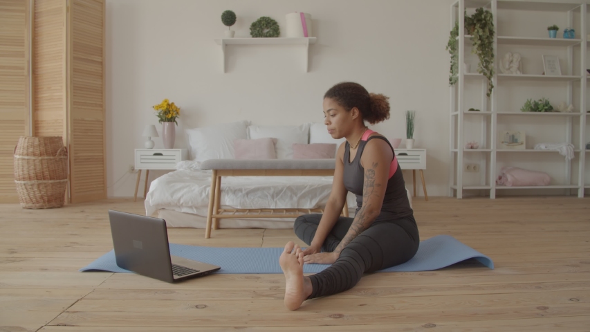 Determined sporty african american woman working out, doing stretching exercise on yoga mat while watching fitness video online on laptop at home. Female practicing yoga exercise online with laptop pc Royalty-Free Stock Footage #1039731266
