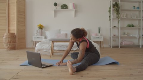 Determined sporty african american woman working out, doing stretching exercise on yoga mat while watching fitness video online on laptop at home. Female practicing yoga exercise online with laptop pc