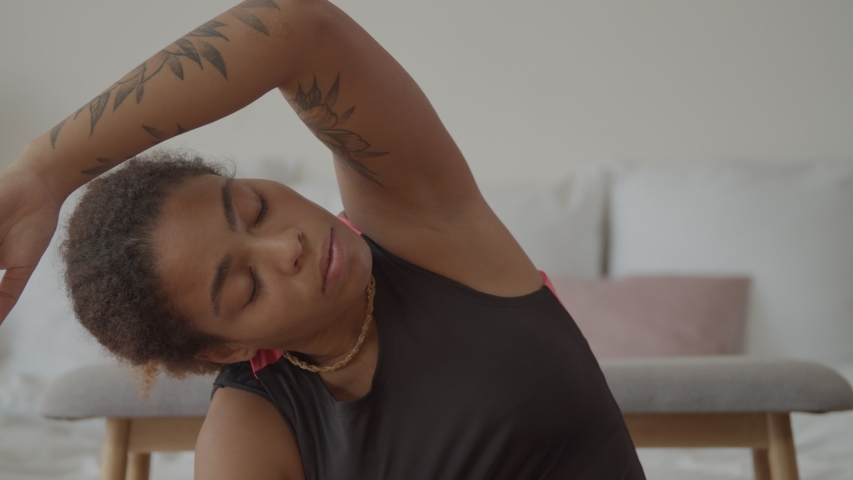 Portrait of relaxed pretty black woman with eyes closed stretching arms over head during indoor workout session. Lovely african american female practicing yoga side bending exercise in domestic room. Royalty-Free Stock Footage #1039731275