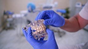 Doctor shows fake implants on a plastic jaw sample or model. Modern dental clinic background. Blue medical gloves on doctor's hands. Dental Hygiene and Health concept video. Implants in jaw.