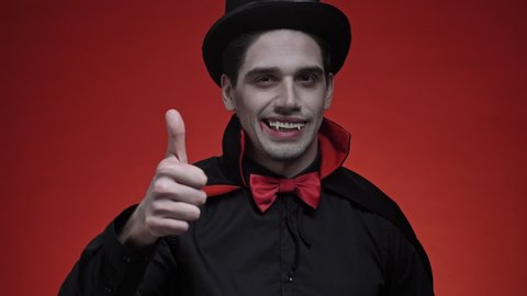 Happy vampire man with blood and fangs in black halloween costume showing thumb up isolated over red wall