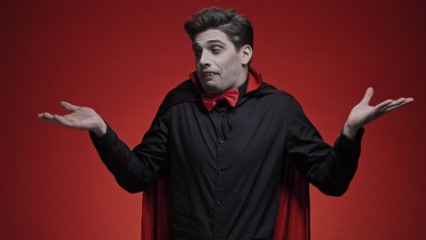 Vampire man with fangs in black halloween costume being at a loss, showing helpless, mouth curved as if he does not know what to do isolated over red wall