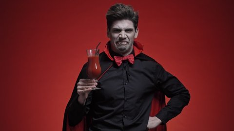 Scary vampire man with fangs in black halloween costume tasting a tomato coctail than disliked it isolated over red wall