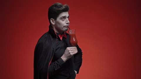 Glad vampire man with fangs in black halloween costume holding a tomato juice and drinking it with a
cocktail tube isolated over red wall