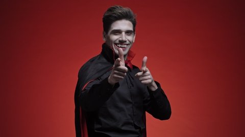 Pleased dracula man with blood and fangs in black halloween costume pointing finger at camera isolated over red wall