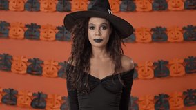 Calm cute young witch woman in black halloween costume yawning and feeling sleepy over orange pumpkin wall