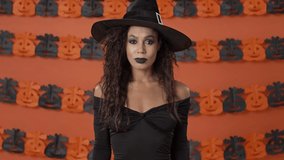 Displeased pretty young witch woman in black halloween costume making thumb down gesture and shaking her head negatively over orange pumpkin wall