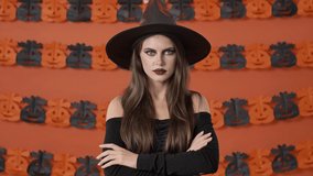 Serious pretty young witch woman in black halloween costume saying no and shaking her head negatively over orange pumpkin wall
