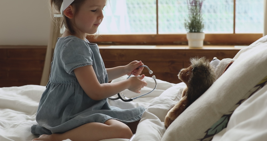 Preschool kid girl pretending doctor listening treating toy patient in bedroom, cute funny little child wear nurse medical hat playing game with stethoscope helping fluffy hedgehog in bed at home Royalty-Free Stock Footage #1039756256
