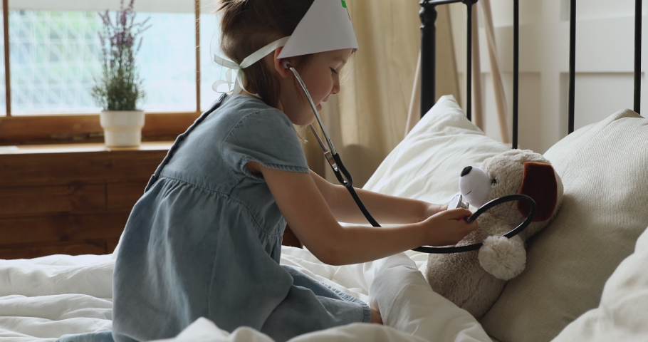 Cute little kid girl playing funny game as vet doctor hold stethoscope listening toy in bedroom, funny preschool child wear medical hat pretending nurse treat fluffy dog patient lying in bed at home Royalty-Free Stock Footage #1039756262