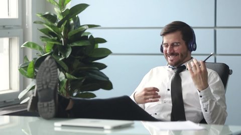 Businessman resting in office room workplace put legs on table leaned on chair wearing headphones imitating playing a guitar enjoy listen music during break, stress relief, hobby or relaxation concept