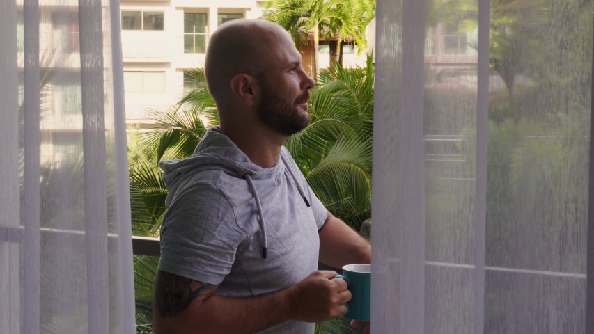 An adult man is drinking coffee on the balcony of the hotel. He is looking at palm trees. Vacation time