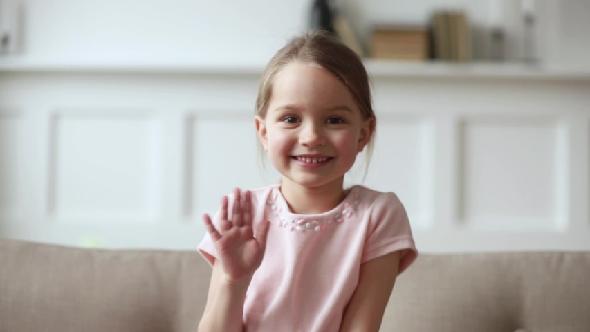Little 6 years old cutie girl sitting on couch wave hand greeting friend or relative person feels shy, answering questions using online telecommunications application video call virtual talk concept Royalty-Free Stock Footage #1039782809