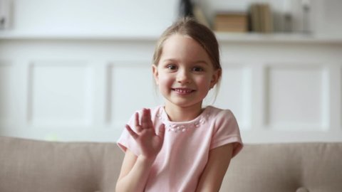 Little 6 years old cutie girl sitting on couch wave hand greeting friend or relative person feels shy, answering questions using online telecommunications application video call virtual talk concept