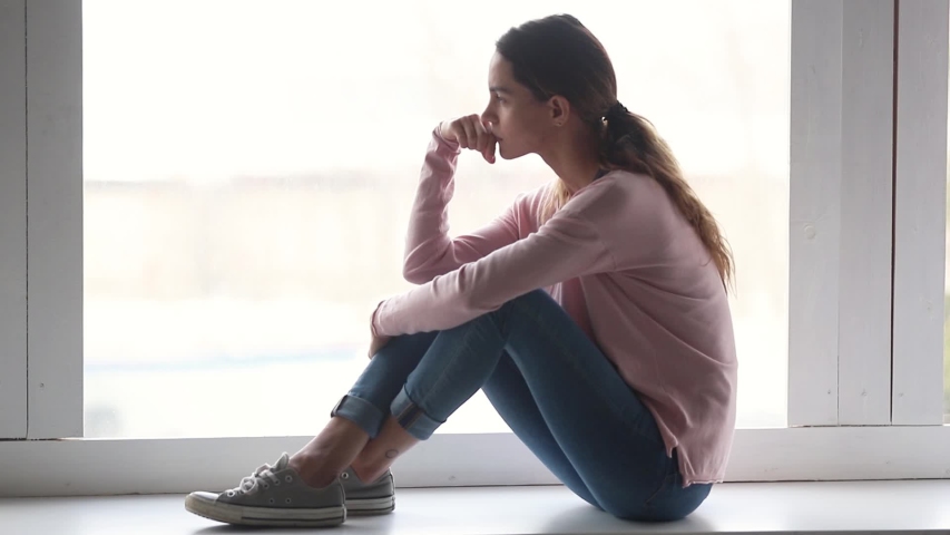 Side view on window background pensive sad young woman sitting on windowsill looks away and thinking having melancholic mood, or personal problems. Break up, loneliness and unrequited love concept | Shutterstock HD Video #1039785890