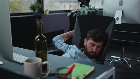 Drunk exhausted young man falling asleep in chair at office table. Stupid-looking fun business manager relaxing in chair falling on ground. Alcohol addiction.