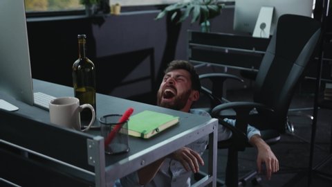 Fun office worker going crazy of alcohol lying on the floor and laughing hard. Drunk addicted business manager getting up sitting in chair. Alcohol intoxication.