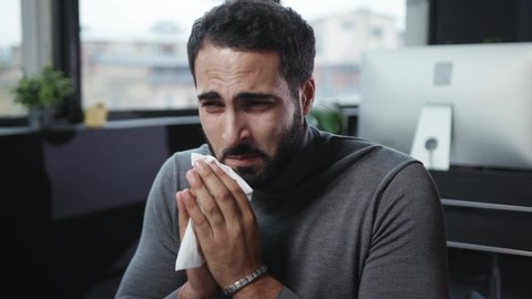 Exhausted caucasian business manager coughing suffering cold and allergy blowing his nose with paper tissues trying to work on desktop computer in the office.