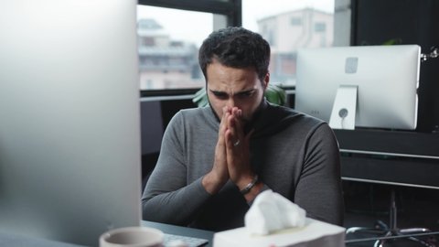 Handsome caucasian man working on personal computer in contemporary office. Sick business employee sneezing allergy suffering cold using napkins at table.