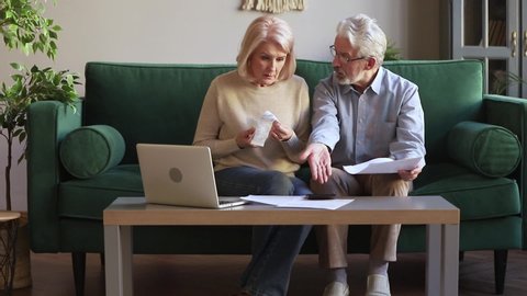 Stressed elderly spouses couple sit on couch in living room managing family budget checking expenses feels anxiety worried about overdue, high taxes, lack deficit of money accounting problems concept
