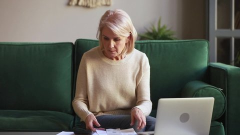 Elderly concentrated european woman sitting on couch at home manages personal family budget counts money holding cheques calculates domestic bills using calculator online banking on pc feels worried