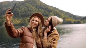 two funny attractive girls young women fashionably dressed on vacation on a lake in the mountains take a selfie on the mobile phone. lesbian love. concept, advertising, friends