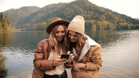 two funny attractive girls young women fashionably dressed on vacation on a lake in the mountains look into a mobile phone and laugh