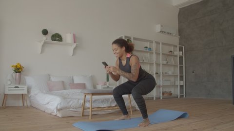 Multitasking sporty fitness african american woman doing squats and browsing online on smart phone in loft apartment. Busy with cellphone fit attractive black female working out and squatting at home.