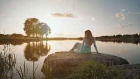 A lonely girl sits on a lake on a stone on a sunset background. Video on the move.