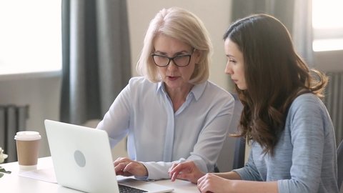 Aged mentor helping with enterprise program on computer to intern young inexperienced girl, businesswomen different generations ages working together on task or project sitting at desk in workplace