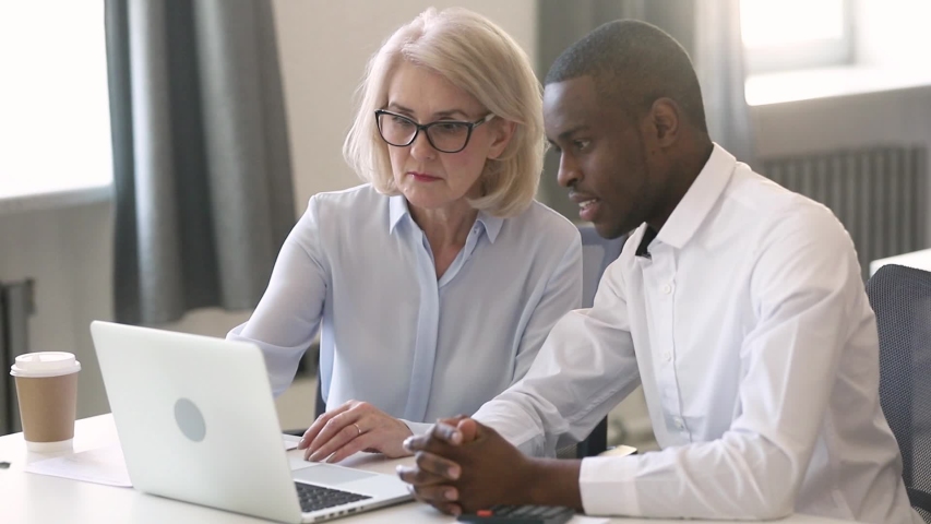 Different age and ethnicity office workers sitting at desk look at laptop screen, leader of project aged businesswoman in glasses explain details to african employee intern inexperienced man colleague Royalty-Free Stock Footage #1039793729