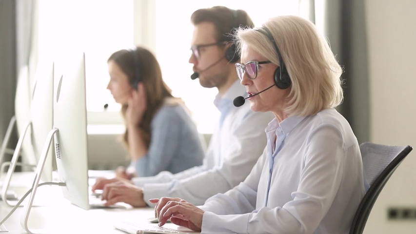Aged woman call center employee sitting at workplace having busy workday, sales agent wearing headset use computer typing search information provide help to client distantly helpline office concept Royalty-Free Stock Footage #1039793774