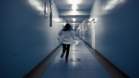Tracking shot of zombie woman with black hair in laboratory coat walks down dark corridor goes behind door and falls,view from back. Above doorway inscription in Russian exit.