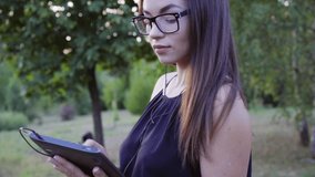 the girl in glasses walks in the Park and looks at the tablet, talking to friends and family.