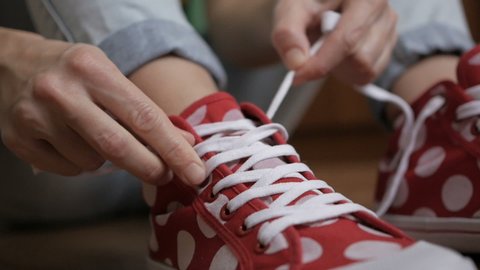 Medium shot. Woman tying shoelaces of red sneaker sitting on a floor. Closeup on girl hands tying laces ready for sport, walking street. Slow motion. 