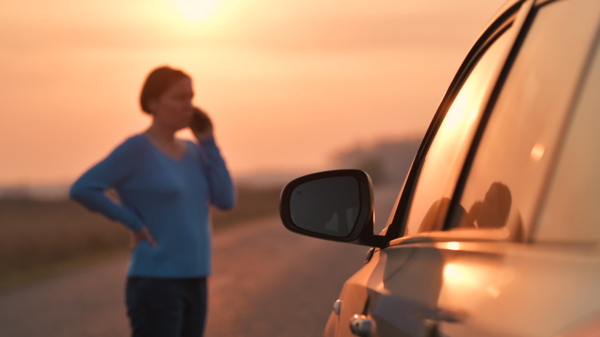 Woman calling on mobile phone for assistance and help on the road after her car broke down in autumn sunset on road through countryside, selective focus
