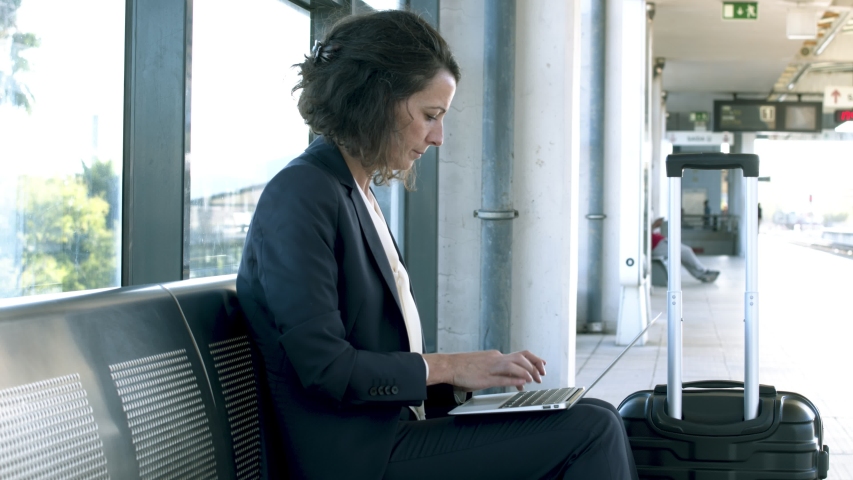 Businesswoman working with laptop at railway station. Side view of middle aged business woman sitting with luggage at railway station and having video chat via laptop. Technology concept