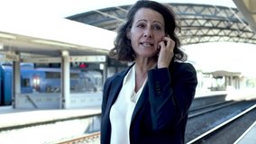 Businesswoman with mobile phone at train station. Smiling middle aged businesswoman in formal wear talking by smartphone at railway station. Technology concept