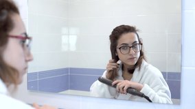 A young woman in glasses straightens her hair with a curling iron in front of a mirror. A girl in a white coat makes a hairstyle in the bathroom. View through the mirror