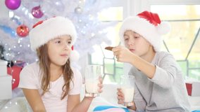 Cute little kids in Santa hats drinking milk and eating delicious cookies at home, looking fully satisfied.