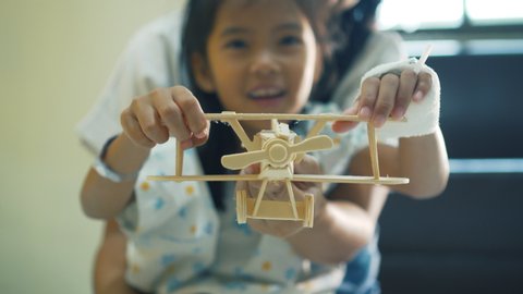 Sick asian little girl playing toy wooden airplane with her mother together while stay in the hospital bed. Therapy for patients to relax. Recovering little girl.