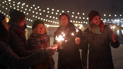Group of friends having fun waving with sparklers in hands. Happy people partying on winter night with snowfall, Christmas market and lamp garlands on background. New Year Holidays or Birthday party. Arkivvideo