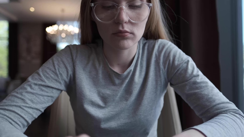 A young woman sits with a pen in her hands at a table near a large window and thinks