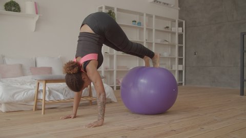 Motivated pretty african american woman in sports clothes practicing fitball pike jackknife push-ups combo, exercise for upper body, abs, back and lower body during intense workout in domestic room.