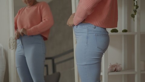 Upset overweight african american woman buttoning old tight pants with difficulties after gaining weight. Frustrated black female struggling to button small jeans in front of mirror at home.