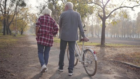 Mature Caucasian couple walking with bicycle along the alley in the park. Happy retired senior family spending autumn evening outdoors. Aging together, eternal love concept.