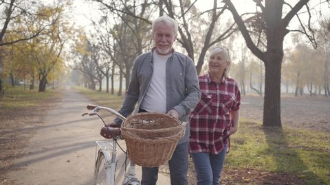 Happy senior Caucasian couple walking with bicycle along the alley in the foggy park. Retired mature family spending autumn evening outdoors. Aging together, eternal love concept.