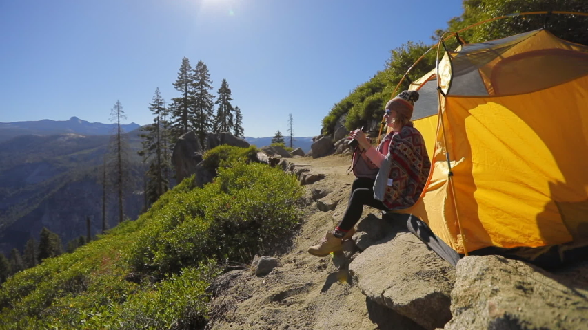 The view of the Yosemite Valley from a mountain path on a hillside.A yellow tent stands on the trail. A girl sits at the edge of a tent and watches the valley through binoculars.Yosemite National Park
