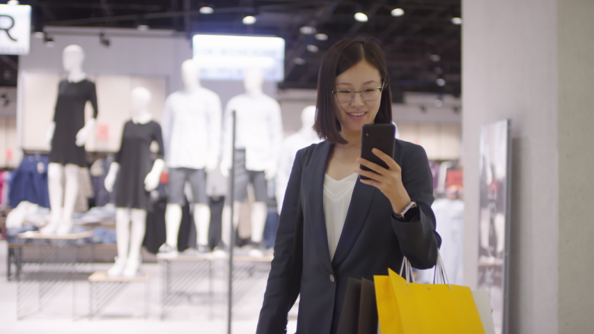 Tracking and lockdown shot of smiling young Chinese woman with plenty of paper bags on hand walking through shopping mall past clothing store and texting on smartphone | Shutterstock HD Video #1039889945